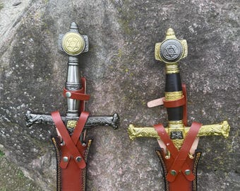May include: Two decorative swords with ornate hilts and crossguards are displayed. The hilts feature a gold and silver design with a star emblem. The swords are sheathed in brown leather scabbards with decorative stitching.