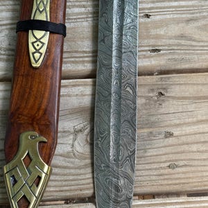 May include: A Damascus steel blade with a distinct wavy pattern alongside a wooden scabbard. The scabbard features a brass fitting with an eagle motif at the tip and a decorative brass accent secured by two black bands near the top section.