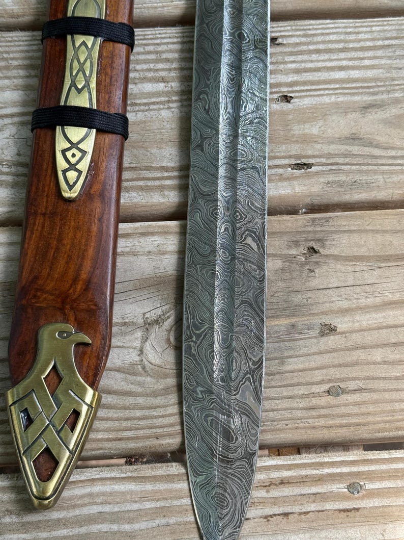 May include: A Damascus steel blade with a distinct wavy pattern alongside a wooden scabbard. The scabbard features a brass fitting with an eagle motif at the tip and a decorative brass accent secured by two black bands near the top section.