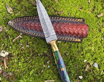 Può includere: Un coltello in acciaio Damasco con una ghiera in ottone e un manico marmorizzato colorato. Il coltello poggia su un fodero in pelle marrone con un disegno intrecciato, su uno sfondo di muschio verde.