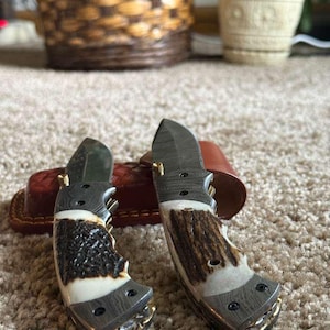 May include: Two folding knives with textured, antler-like handles and dark blades. The handles feature a combination of brown, white, and dark grey tones. The knives are resting on a brown leather sheath.