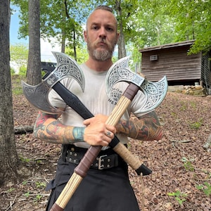 May include: Two silver-bladed axes with intricate designs are crossed in front of a person. One axe has a black handle, the other a light brown handle. The person is wearing a light-colored shirt and a black kilt. The axes are displayed outdoors.