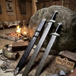 May include: Two swords with silver blades and black handles are displayed with a black sheath. The swords are placed on a fur-like surface, alongside a wooden box, a drinking horn, and a sign that reads "SKALDIK EDGE EST. 2024".