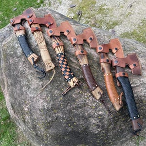 May include: Six Viking-style axes with leather sheaths and handles. The axe heads are made of metal, and the handles are wrapped in leather or wood. The axes are displayed on a large gray rock, with a grassy background.