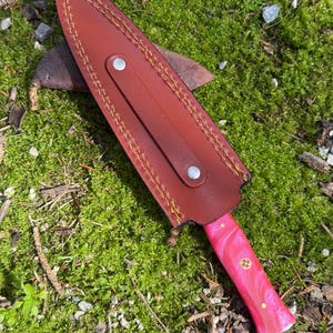Pu&ograve; includere: Un coltello con manico rosa e rosso con un fodero in pelle marrone. L'impugnatura del coltello ha un emblema dorato decorativo. Il fodero &egrave; cucito con filo giallo e ha un cinturino con un bottone argentato.