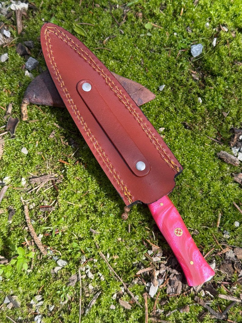 Pu&ograve; includere: Un coltello con manico rosa e rosso con un fodero in pelle marrone. L'impugnatura del coltello ha un emblema dorato decorativo. Il fodero &egrave; cucito con filo giallo e ha un cinturino con un bottone argentato.