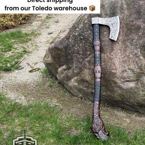 May include: A Viking-style axe with a silver blade featuring intricate Celtic knotwork. The handle is wrapped in black and brown leather, with a carved wooden base. The axe rests against a large rock on a grassy surface.