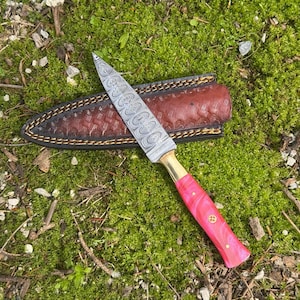 Pu&ograve; includere: Un coltello in acciaio Damasco con manico rosa e ghiera in ottone poggia su un fodero in pelle marrone. La lama presenta un motivo unico. Il coltello &egrave; appoggiato su uno sfondo di muschio verde.