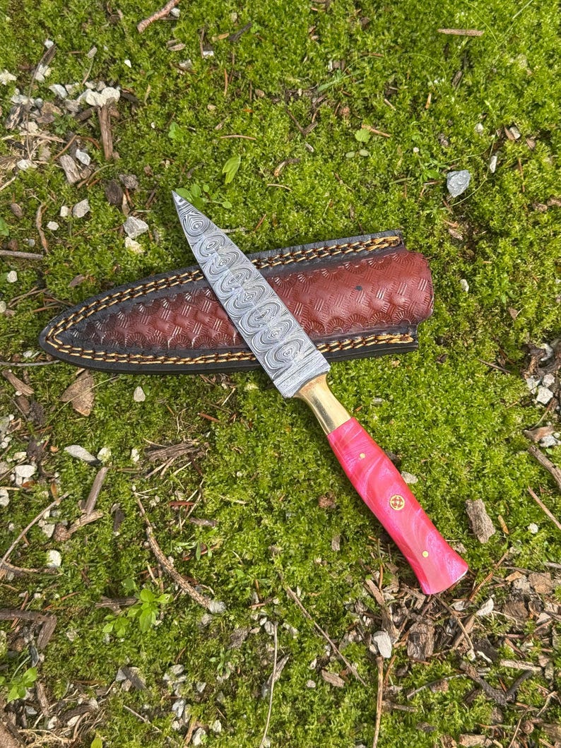 Pu&ograve; includere: Un coltello in acciaio Damasco con manico rosa e ghiera in ottone poggia su un fodero in pelle marrone. La lama presenta un motivo unico. Il coltello &egrave; appoggiato su uno sfondo di muschio verde.
