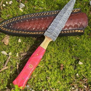 Pu&ograve; includere: Un coltello fatto a mano con una lama in acciaio fantasia e un manico rosa. Il coltello ha una ghiera in ottone ed &egrave; accompagnato da un fodero in pelle marrone con cuciture decorative. Il coltello &egrave; appoggiato su muschio verde.