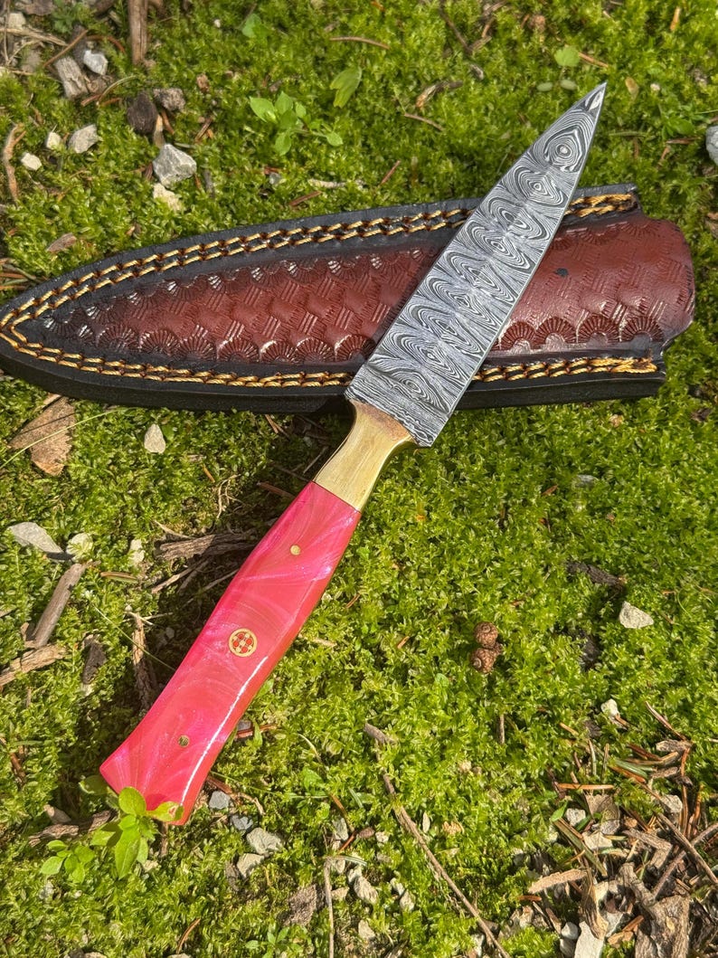 Pu&ograve; includere: Un coltello fatto a mano con una lama in acciaio fantasia e un manico rosa. Il coltello ha una ghiera in ottone ed &egrave; accompagnato da un fodero in pelle marrone con cuciture decorative. Il coltello &egrave; appoggiato su muschio verde.
