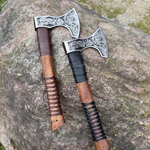 Pode incluir: Dois machados vikings com cabos de madeira e cabeças de machado prateadas com designs intrincados. Um machado tem um cabo envolto em couro marrom, e o outro tem um cabo envolto em couro preto. Os machados são exibidos em uma grande rocha cinza.