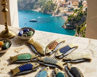 Può includere: Una collezione di coltelli tascabili decorativi a forma di pesce con dettagli dorati e squame colorate, tra cui blu, verde e marrone. I coltelli sono disposti su una superficie di marmo, con una città costiera italiana sullo sfondo.
