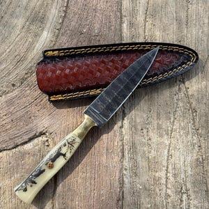 May include: A handcrafted knife with a patterned blade and a cream-colored handle featuring a tree design. The knife has a brass bolster and rests on a wooden surface next to a brown leather sheath with decorative stitching.