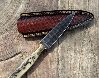Può includere: Un coltello artigianale con una lama a fantasia e un manico color crema con un disegno ad albero. Il coltello ha una ghiera in ottone e poggia su una superficie di legno accanto a un fodero in pelle marrone con cuciture decorative.