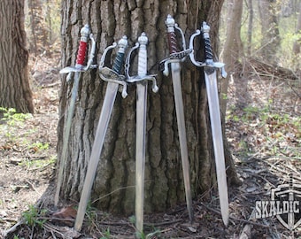 May include: Five swords with silver blades and ornate handles lean against a tree. The handles are wrapped in red, teal, white, brown, and black. The swords are displayed outdoors in a wooded area.