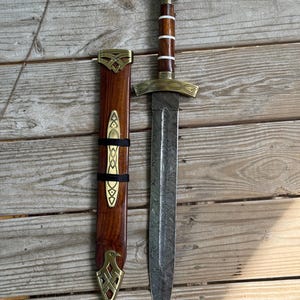 May include: A double-edged sword with a patterned Damascus steel blade, a wooden handle with white bands, and brass fittings. The matching wooden scabbard features brass accents, including a decorative locket, a central plate, and a bird-shaped chape.
