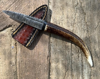 Può includere: Un coltello artigianale con una lama strutturata e un manico in corno. Il coltello è in un fodero di cuoio marrone con cuciture decorative. La lama misura circa 10 cm.