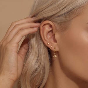 Könnte beinhalten: Nahaufnahme eines Ohrs mit drei goldenen Ohrringen. Ein kleiner Stecker, ein kleiner Reifen und ein Reifen mit einem Sternanhänger. Die Ohrringe sind vor blondem Haar und Haut zu sehen. Das Bild zeigt das Schmuckdesign.