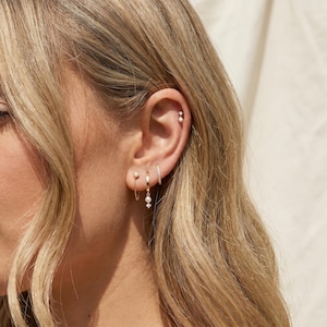 May include: A close-up of an ear adorned with multiple gold earrings. The earrings include small studs, a hoop with a chain and pearl drop, and a small hoop with a row of small crystals. The ear is set against a soft, neutral background.