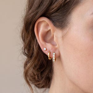 May include: A close-up of a person's ear adorned with three different gold earrings. The earrings include a small stud, a small hoop, and a larger hoop with white opal stones. The earrings are set against the backdrop of the person's hair.
