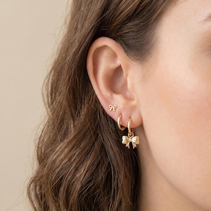 Puede incluir: Pendientes dorados con diseño de lazo. El conjunto incluye pequeños pendientes de botón y aros con un dije de lazo. Los pendientes se muestran en una oreja, mostrando sus delicados detalles y acabado brillante.
