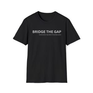 Puede incluir: Camiseta negra con el texto blanco "BRIDGE THE GAP", con el texto más pequeño "(construction may take 3-5 business years)" debajo. La camiseta tiene mangas cortas y cuello redondo.