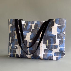 Könnte beinhalten: Eine rechteckige Tragetasche mit einem sich wiederholenden Muster aus dunkelblauen und schwarzen abgerundeten geometrischen Formen auf hellcremefarbenem Grund. Die Tasche hat zwei schwarze Stoffträger, die an der Oberkante befestigt sind.