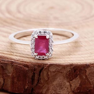 Peut inclure: Une bague en argent ornée d'une pierre précieuse rectangulaire rouge rubis, entourée d'un halo de petites pierres claires. La bague est posée sur un fond en bois, mettant en valeur le design et la couleur du bijou.