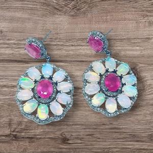 Peut inclure: Une paire de boucles d'oreilles argentées avec une pierre rose ovale centrale et un cercle de pierres opalescentes. Les boucles d'oreilles ont un design floral avec une pierre rose assortie en haut.