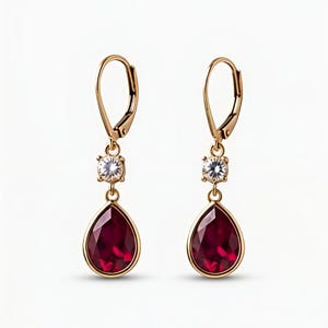 May include: A pair of gold-toned leverback earrings featuring a small, round, clear faceted stone suspended above a larger, pear-shaped, deep red faceted gemstone. The metal settings are polished gold, and the design hangs vertically.