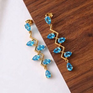 Puede incluir: Un par de pendientes colgantes dorados con un diseño de barra metálica en zigzag. Cada pendiente está adornado con cinco piedras preciosas de color azul claro en forma de pera dispuestas a lo largo del marco, terminando en una gota.