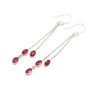 May include: A pair of dangle earrings with three oval-shaped, deep red gemstones each. The gemstones are set in a silver-colored metal and connected by a delicate chain. The earrings have a hook closure.