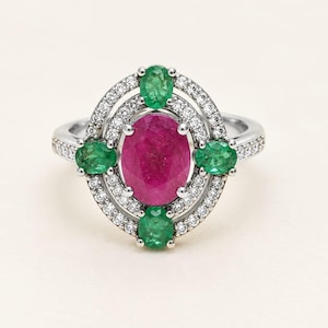 May include: A silver ring featuring a large oval-cut ruby at the center, surrounded by four smaller oval-cut emeralds arranged in a cross pattern. The entire setting is framed by two concentric halos of small, round, clear white diamonds.