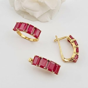 May include: A gold ring and a pair of matching gold hoop earrings, each featuring a row of rectangular, deep red gemstones set in a channel style. The jewellery pieces are arranged together, showcasing the vibrant red stones and polished gold metal.