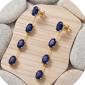 Puede incluir: Un par de pendientes largos de tono dorado con cuatro piedras ovaladas de color azul profundo facetadas, engastadas en garras de metal. Las piedras están conectadas por finas cadenas doradas y los pendientes incluyen cierres de presión estándar.