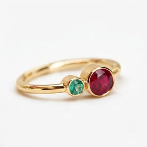 May include: A gold ring featuring two bezel-set gemstones: a smaller round green emerald and a larger round red ruby. The band is smooth and polished, showcasing a minimalist design that highlights the contrast between the two vibrant stones.