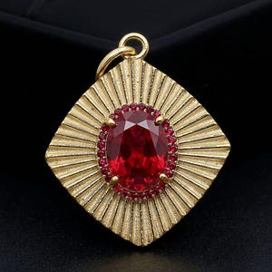 May include: A gold-toned pendant featuring a large, oval-cut deep red central stone surrounded by a halo of small, round red accent stones. The pendant has a square, sunburst-patterned frame with radiating lines and a circular bail at the top.