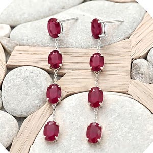 Puede incluir: Un par de pendientes colgantes con cuatro piedras preciosas ovaladas de color rojo intenso y talladas, engastadas en metal plateado. Cada pendiente consiste en una cadena vertical de piedras unidas por pequeños eslabones plateados.