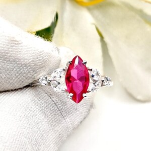 Puede incluir: Un anillo de plata con una gran gema rosa brillante con corte marquesa como pieza central. El anillo está adornado con gemas claras más pequeñas a cada lado, sobre un fondo blanco. El diseño del anillo es elegante y llamativo.