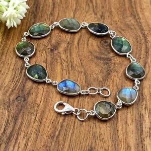 May include: A silver chain bracelet featuring ten faceted, oval-shaped labradorite gemstones. Each stone displays iridescent flashes of blue, green, and grey. The bracelet is secured with a silver lobster claw clasp and includes a small extender chain.