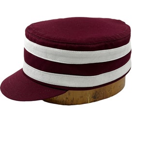 Puede incluir: Una gorra de color granate con una visera blanca y dos rayas blancas alrededor de la corona.