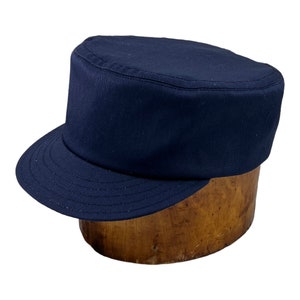 Peut inclure: Une casquette de style militaire bleu foncé avec une visière plate et une couronne rigide. La casquette est faite d'un tissu de coton épais et a un léger brillant.
