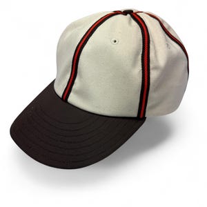 May include: A baseball cap featuring an off-white crown with vertical red and black striped piping along the seams. The cap has a solid dark brown curved brim and includes small ventilation eyelets on the upper panels of the crown.