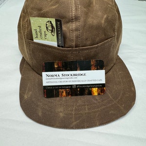 Könnte beinhalten: Braune Fünf-Panel-Kappe mit gebogenem Schirm. Auf der Vorderseite der Kappe befindet sich eine kleine Tasche. Eine Visitenkarte mit dem Text "NORMA STOCKBRIDGE ARTISANAL CREATOR OF INDIVIDUALLY CRAFTED CAPS" befindet sich auf dem Schirm.