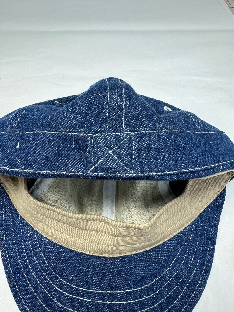 K&ouml;nnte beinhalten: Blaue Jeans-Cap mit wei&szlig;en N&auml;hten. Die Cap hat einen gebogenen Schirm und ein beigefarbenes Innenfutter. Die N&auml;hte bilden ein Kreuzmuster auf der Oberseite.