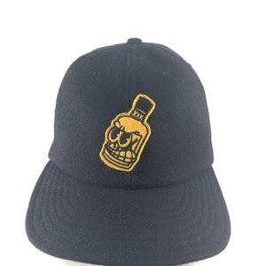 May include: Black baseball cap with a gold embroidered skull wearing sunglasses and a top hat. The skull is holding a bottle with the letters 'XXX' on it.