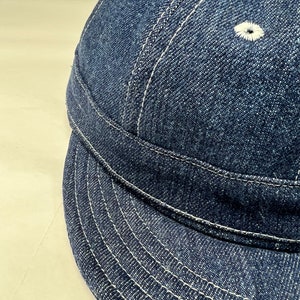 K&ouml;nnte beinhalten: Nahaufnahme einer Baseballkappe aus Jeansstoff. Die Kappe ist dunkelblau mit wei&szlig;en Nahtdetails. Der Schirm ist gebogen und die Kappe hat zwei kleine, wei&szlig; gestickte &Ouml;sen auf der Oberseite.