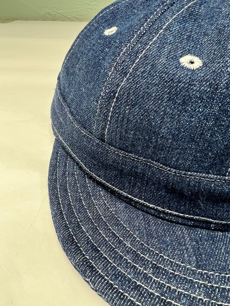 K&ouml;nnte beinhalten: Nahaufnahme einer Baseballkappe aus Jeansstoff. Die Kappe ist dunkelblau mit wei&szlig;en Nahtdetails. Der Schirm ist gebogen und die Kappe hat zwei kleine, wei&szlig; gestickte &Ouml;sen auf der Oberseite.