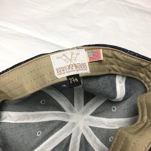 K&ouml;nnte beinhalten: Eine Baseballkappe aus Jeansstoff mit einem beigefarbenen Innenband und wei&szlig;en N&auml;hten. Die Kappe hat ein Etikett mit dem Text "STOCKBRIDGE SEWING WORKS" und eine kleine amerikanische Flagge.